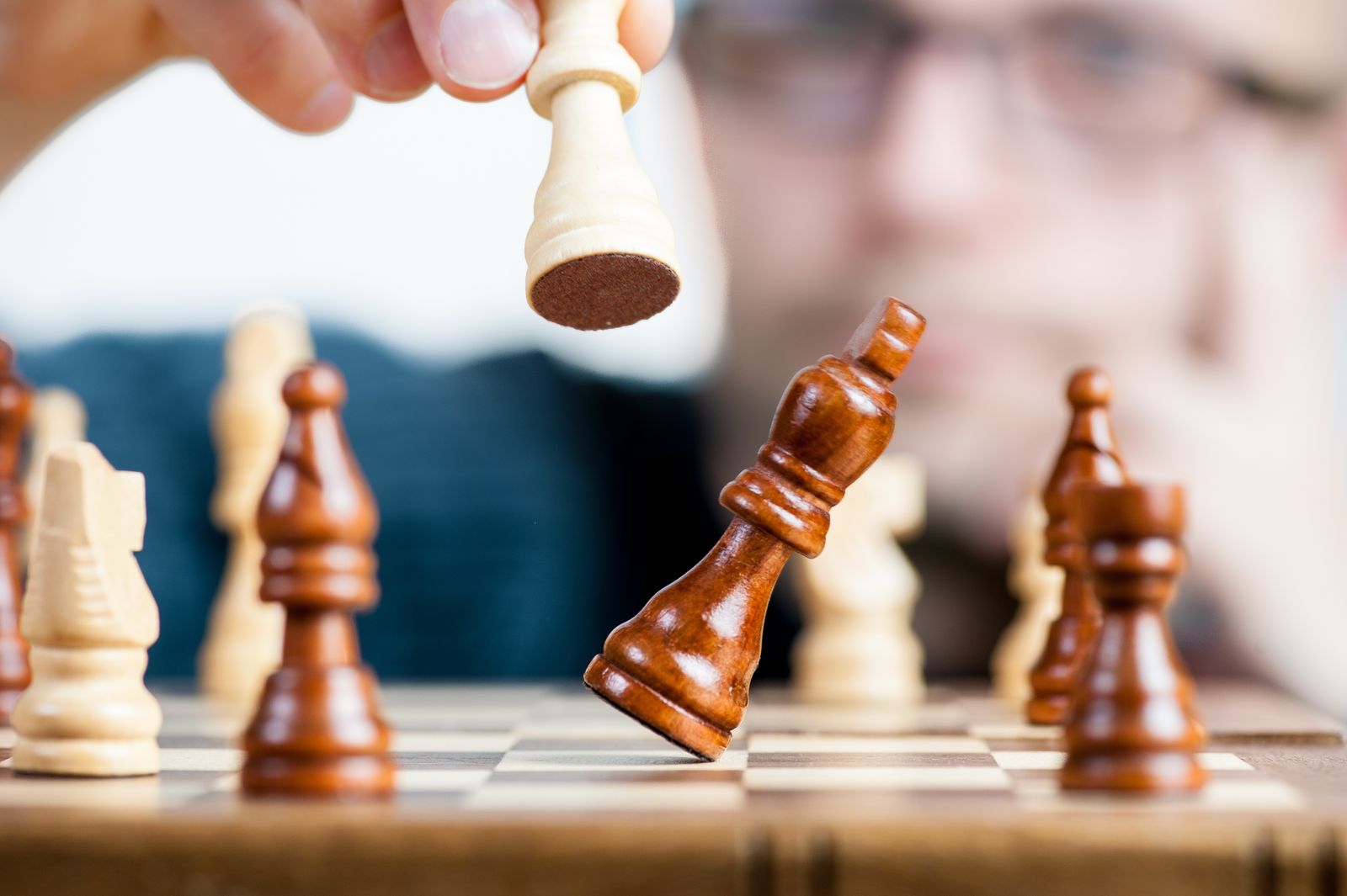 A hand tips over a dark wooden king piece on a chessboard, surrounded by light and dark pieces, the moment a game ends in checkmate or resignation